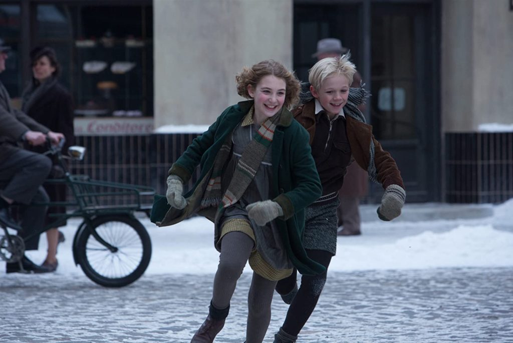 Film still from The Book Thief (2013) of two happy children running in a snowy town square