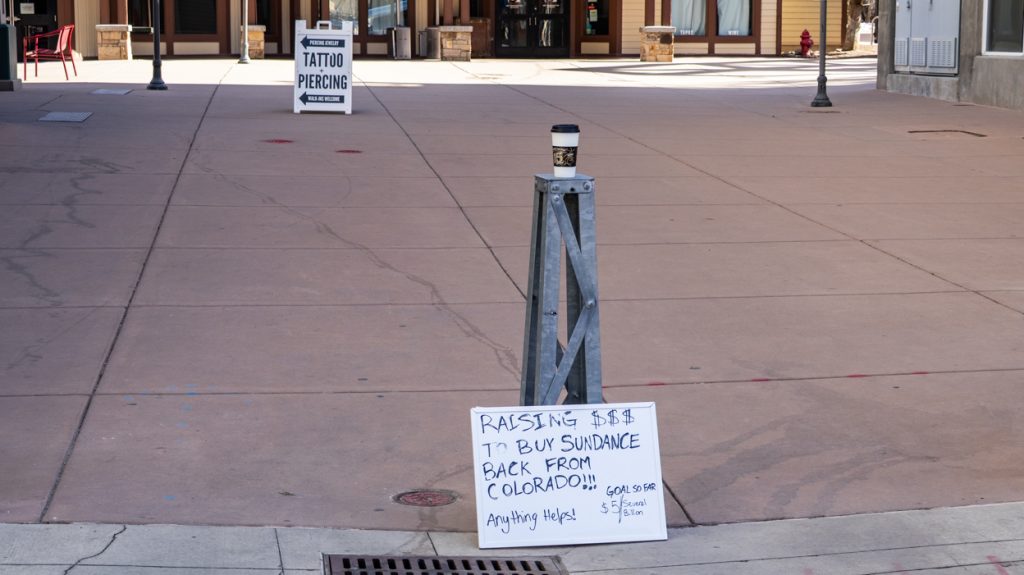 Sign against a bollard stating 'Raising $$$ to buy Sundance back from Colorado!!! Anything helps! Goal so far is $5 out of several billion'
